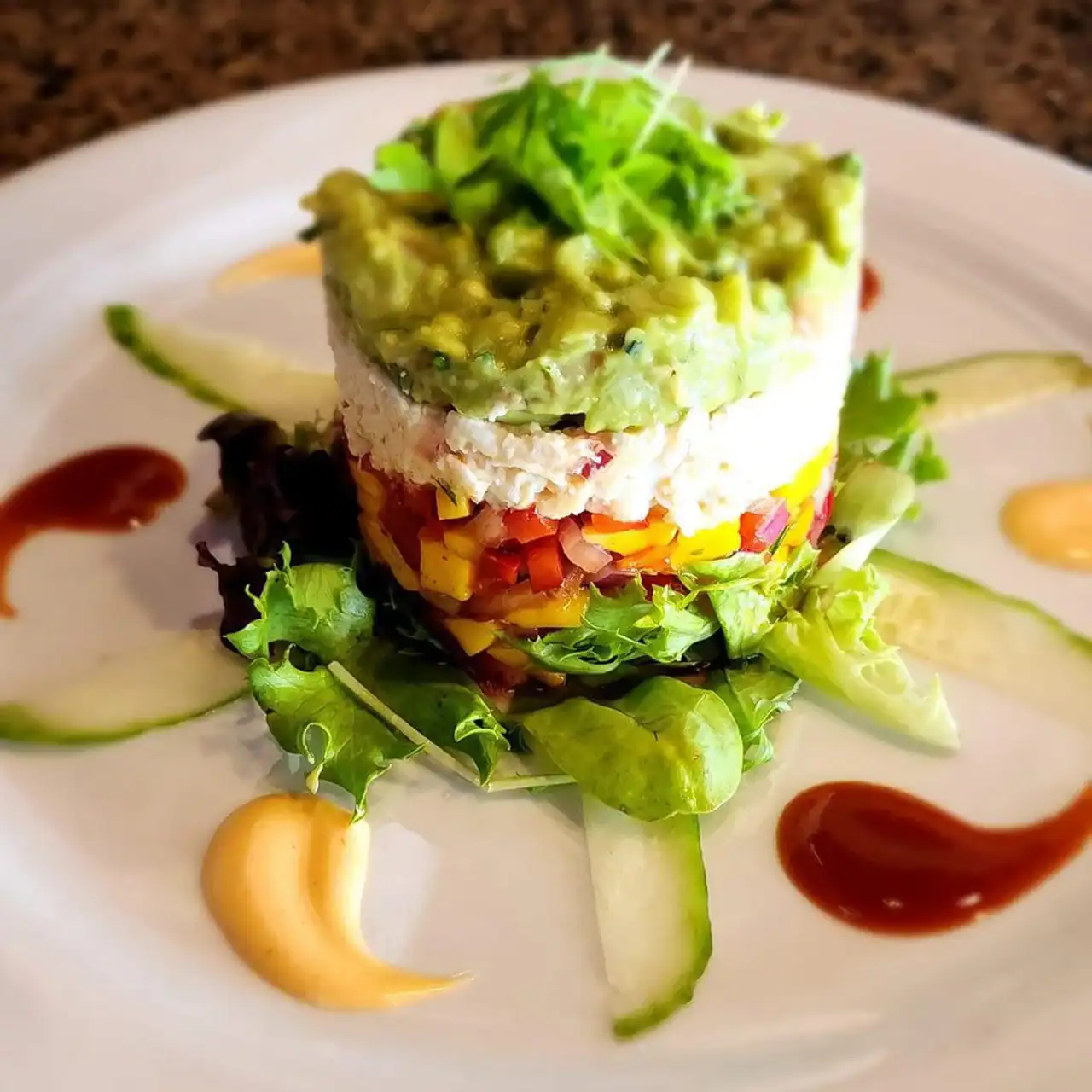 Guacamole and vegetable tower appetizer on a white plate with salad and sauces.