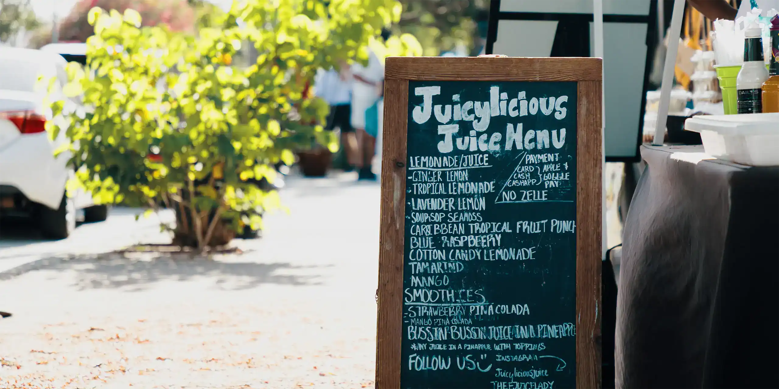 Juicylicious Drink Menu chalkboard sign outdoors at a market or cafe.