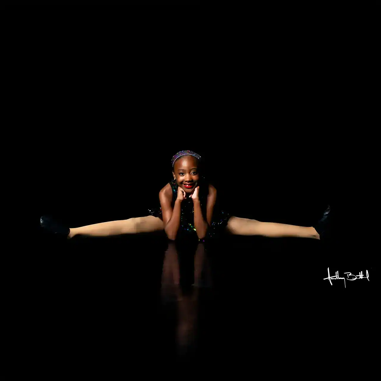A young dancer performs a side split on a reflective black floor, wearing a sparkling outfit and headband.