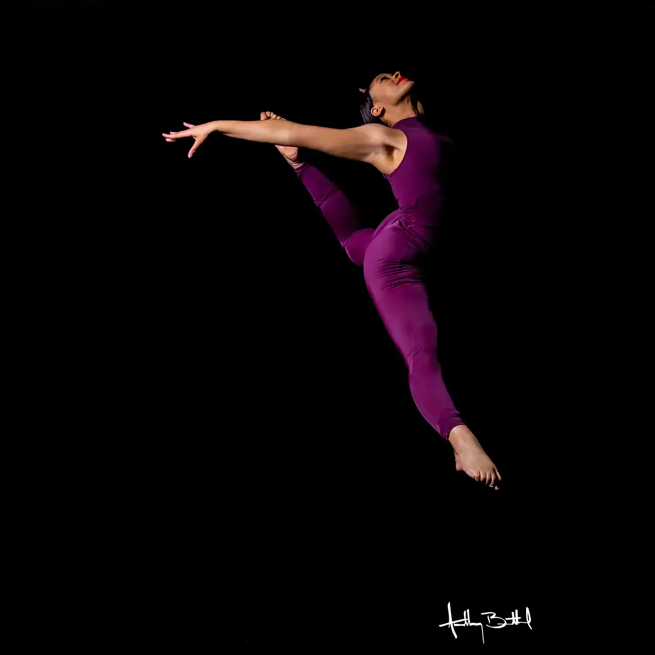 Dance performer in a purple leotard executing a leap against a black background.