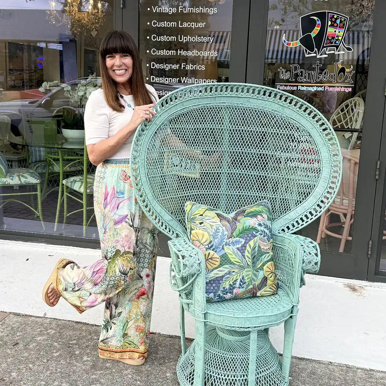 Smiling woman in floral pants poses beside a large, ornate mint-green wicker chair with a colorful leaf-patterned cushion in front of a storefront.