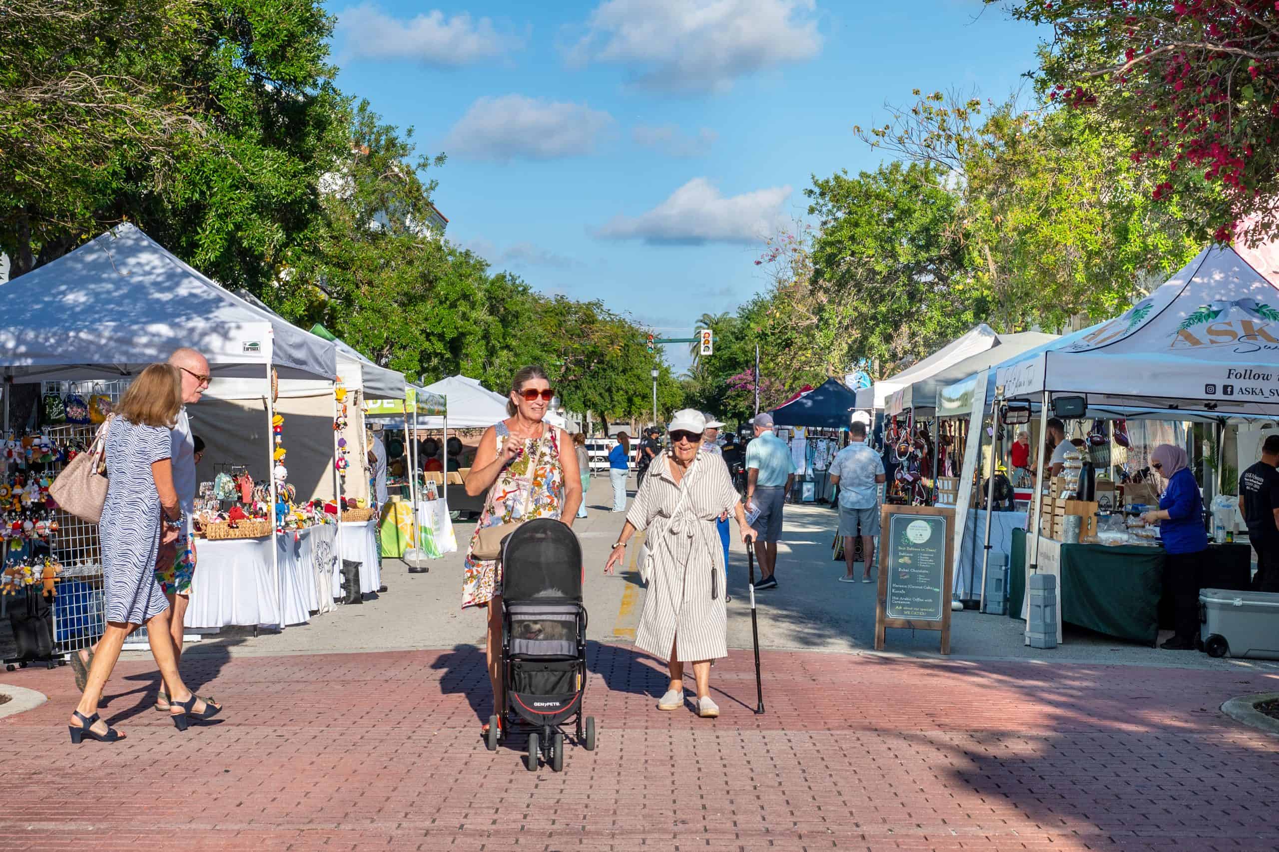 A woman with a stroller and an elderly woman walking through an outdoor market with tents and vendor.