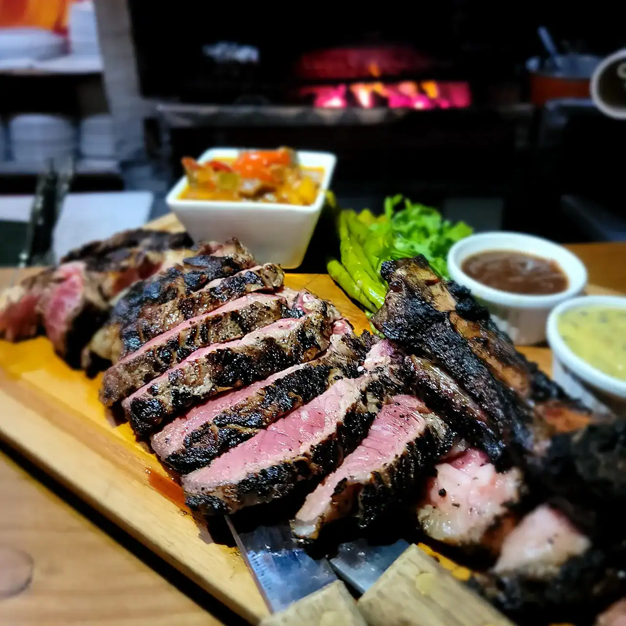 Steak platter with grilled meat, vegetables, and dipping sauces on a wooden board.