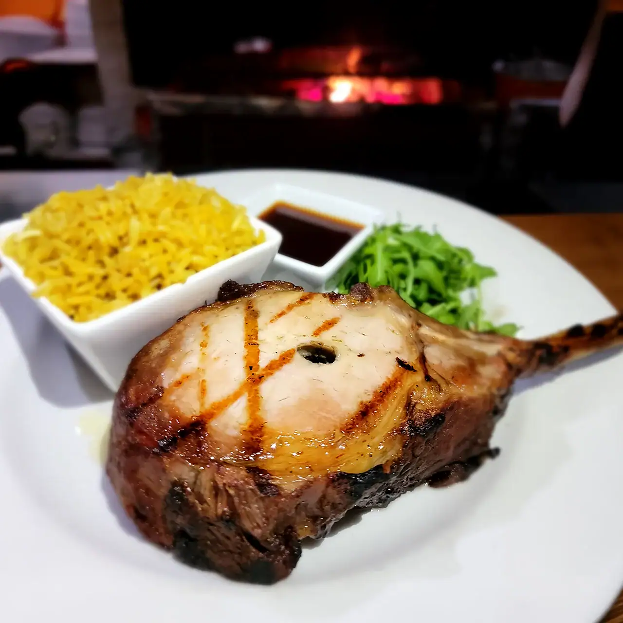 Lamb chop served with yellow rice, fresh greens, and dipping sauce on a white plate.