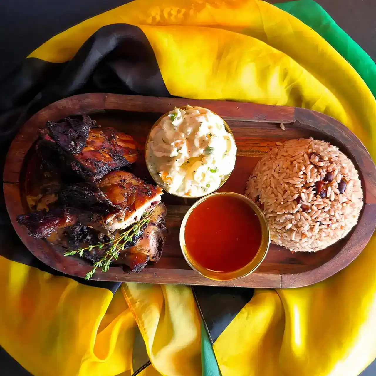 A wooden platter with grilled chicken, a scoop of rice and peas, creamy potato salad, and hot sauce.