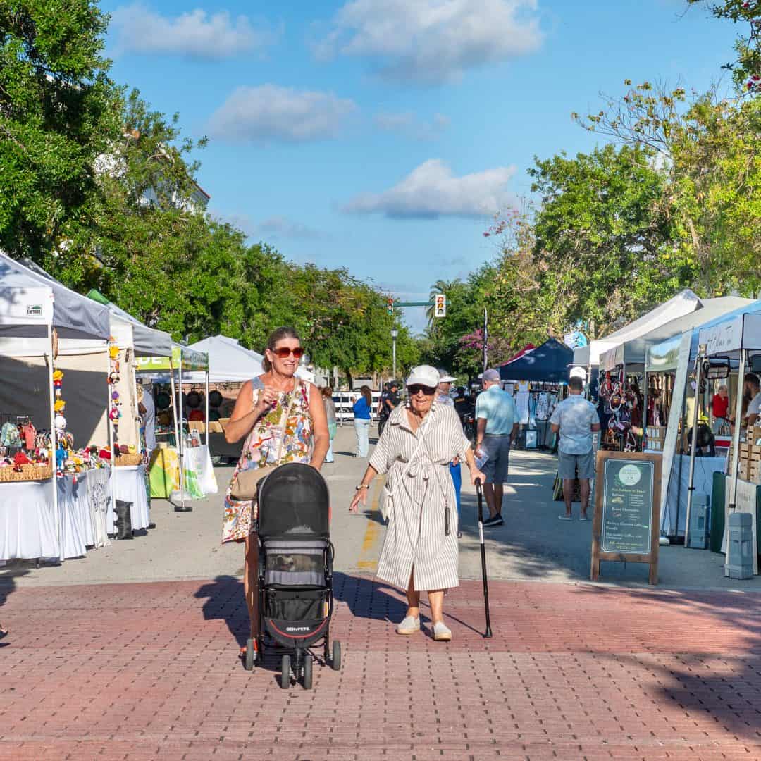 A woman with a stroller and an elderly woman walking through an outdoor market with tents and vendor.